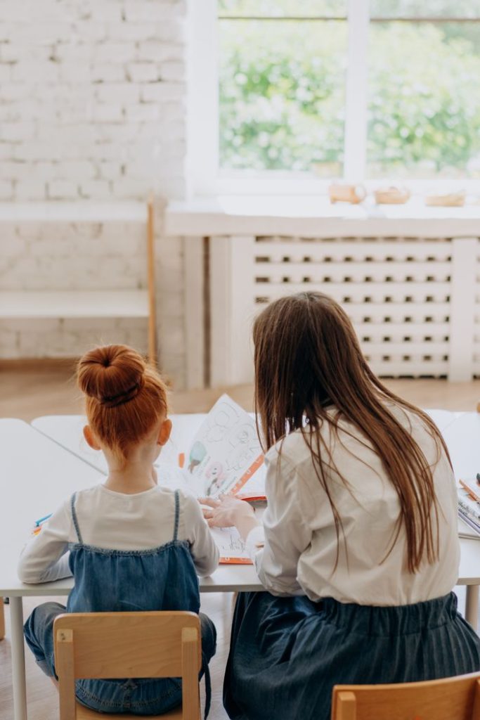 A mother and her daughter sitting at a table, engaged in learning activities inside a bright room.